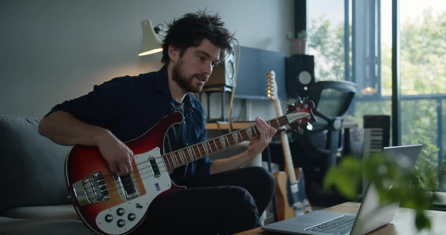 A man talks to a laptop while playing the guitar on his couch