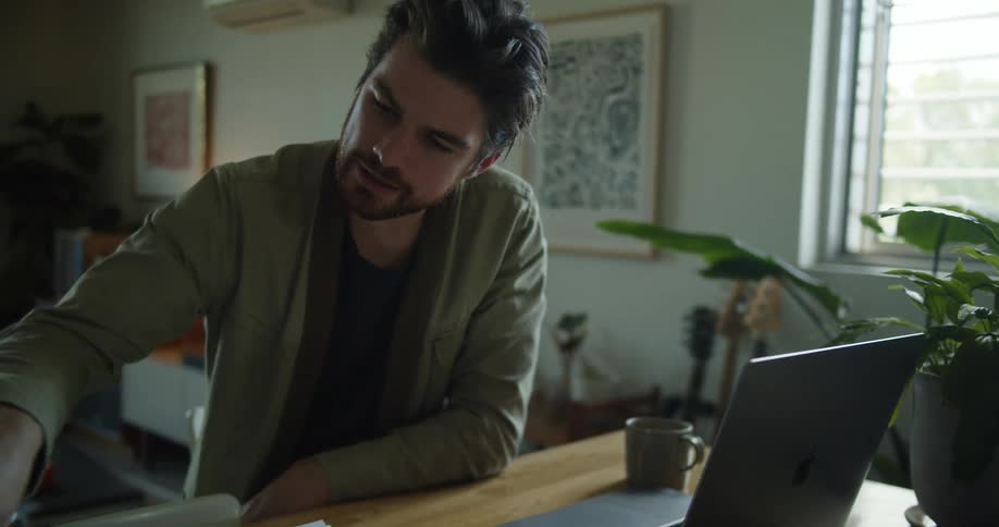 A man sits at a table at home working on a laptop as he flips pages in a binder