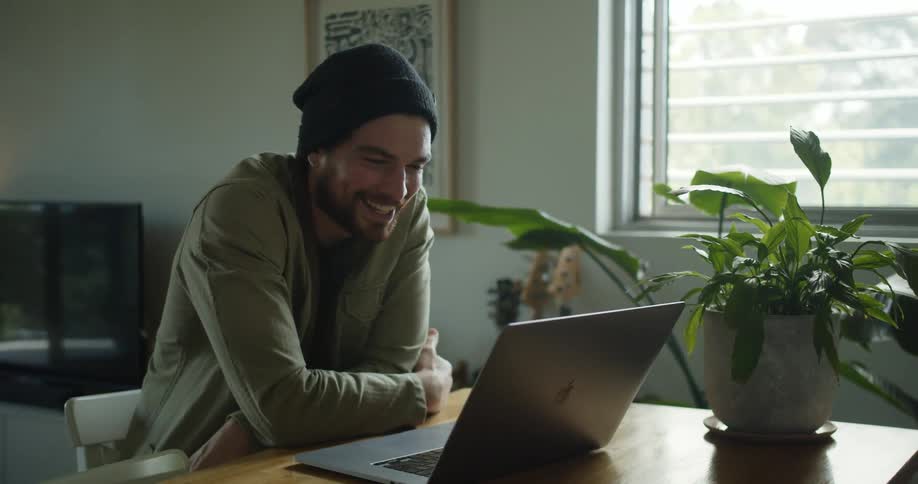 Man with a black beanie and a beard makes a video call on macbook laptop in the living room at home