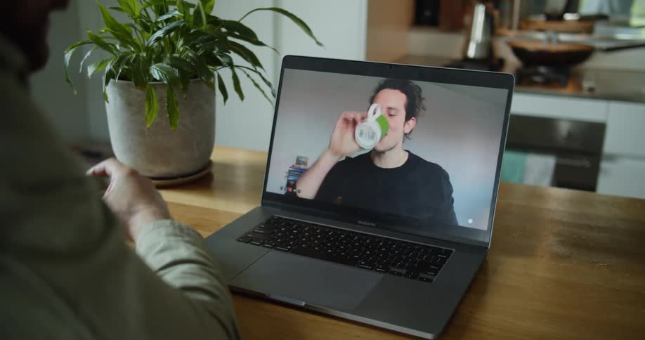 A person sits at a table having a video call with a man and the man drinks from a mug