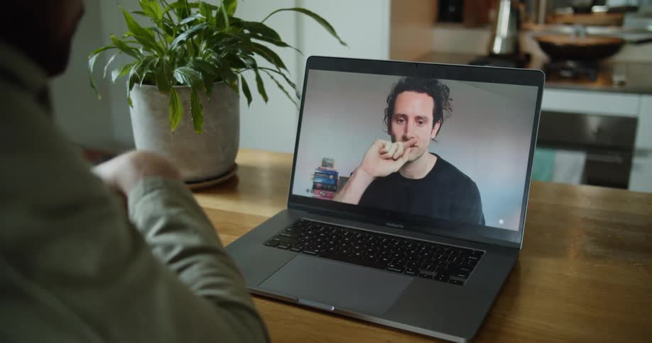 A person sits at a table having a video call with a man on a laptop