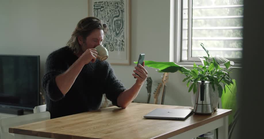 Man with long hair and a black sweater drinks a cup of coffee while making a video call on his iphone smartphone in the living room at home
