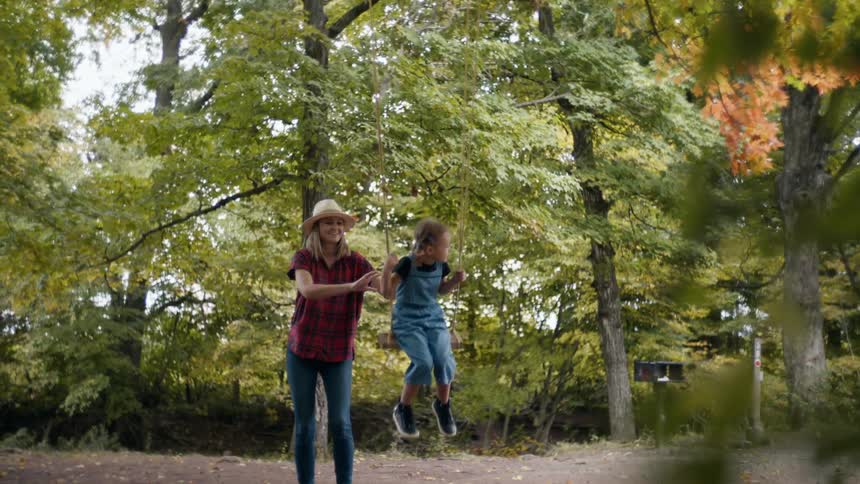 Blonde mother with a hat a red plaid shirt and jeans pushes her daughter on a swing in the woods