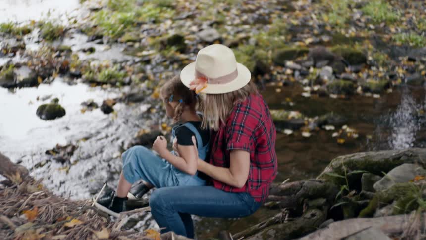 Mother with a hat a plaid shirt and jeans sits on a tree branch and holds her daughter next to the a stream in the woods