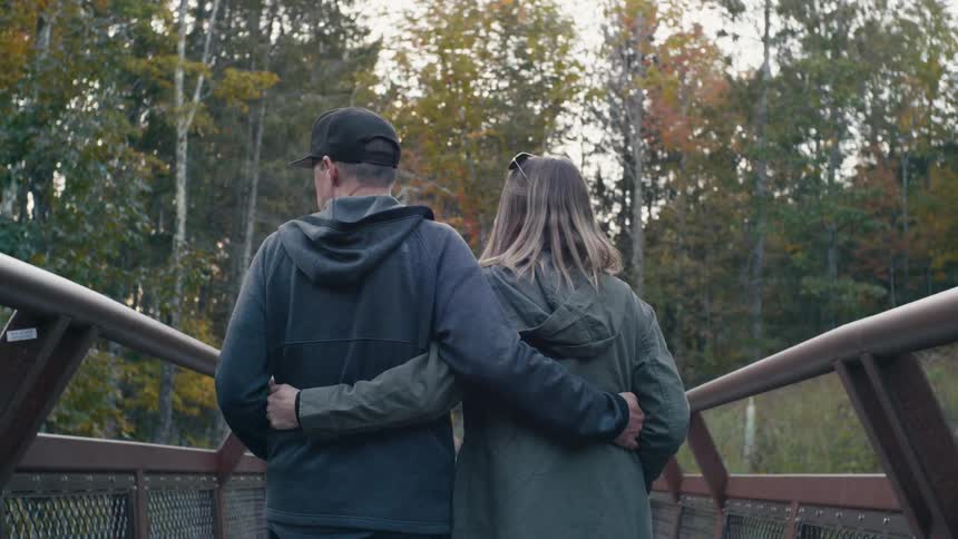 A man and woman holding each other walking across a bridge into a forest