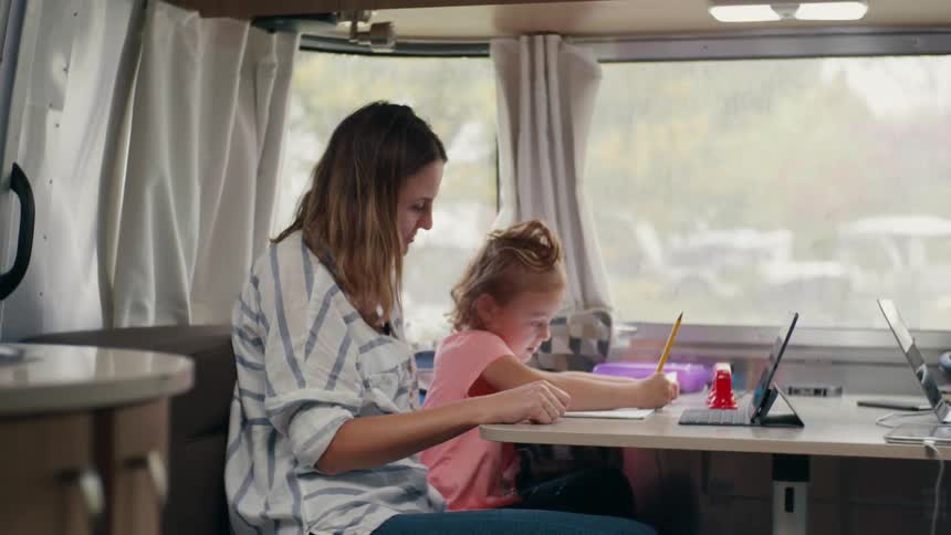 A woman and a girl sitting at a desk with a laptop as the girl draws on a piece of paper with a pencil
