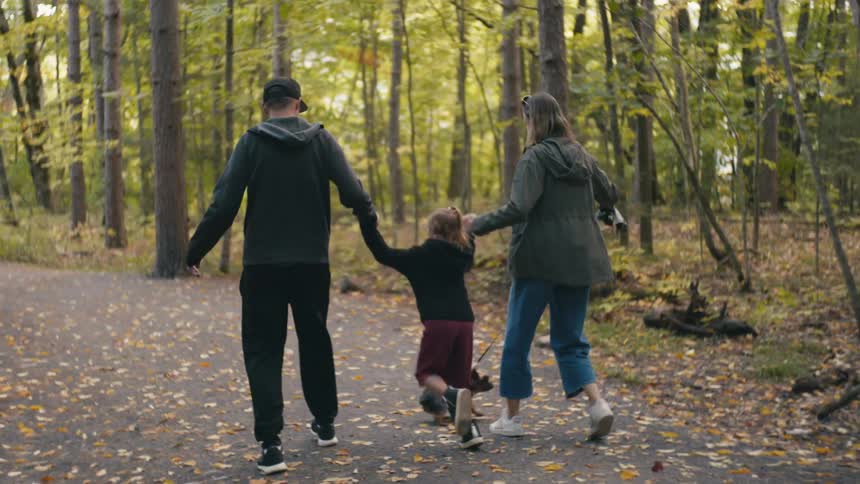 A mom and dad hold hands with their daughter and swing her as they walk on a path in a forest with a dog
