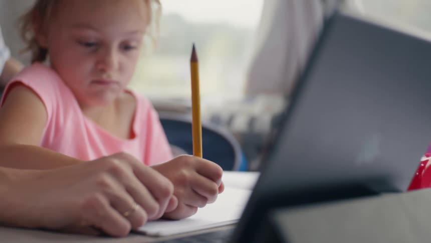 A girl sits in her mothers lap in front of a tablet while doing homework