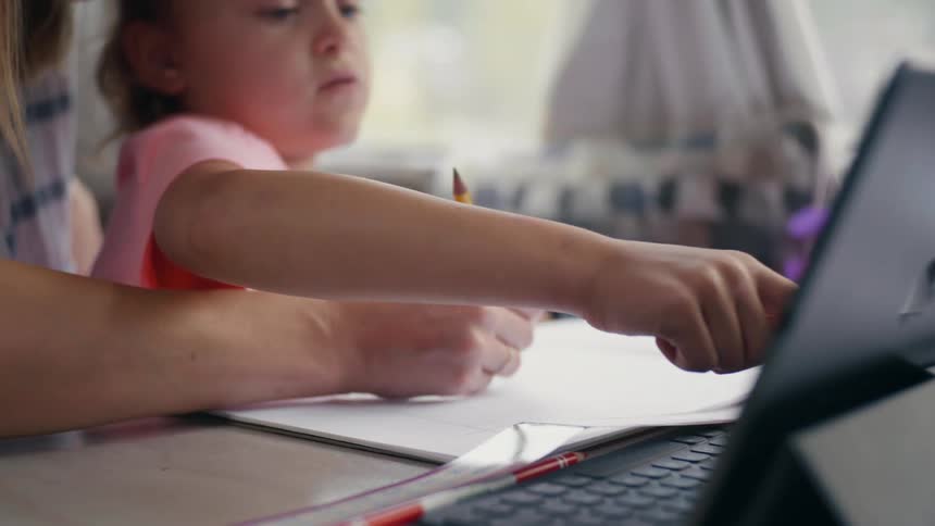 A woman draws on a piece of paper with a toddler girl pushing something on a laptop screen
