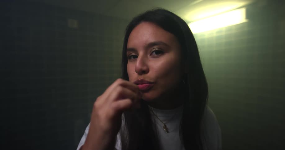 Brunette woman with dark straight hair and a grey sweatshirt brushes her teeth looking at the camera in the bathroom