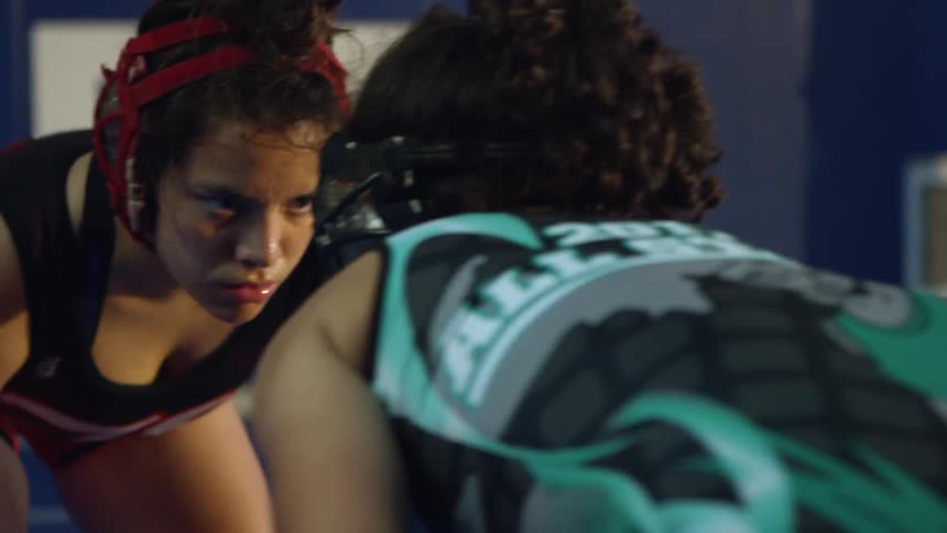 Two girls wearing wrestling gear wrestle indoors