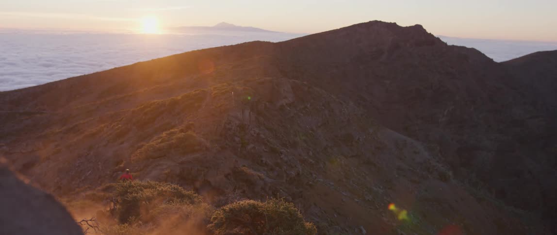 Person rides bike up mountain at sunset with the sea in the distance