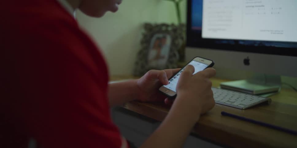 Teenage boy using a smartphone in front of a computer on a desk at home