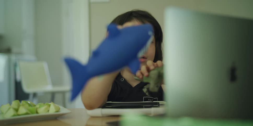 Little boy playing with a toy shark and toy dinosaur in front of a laptop at a table