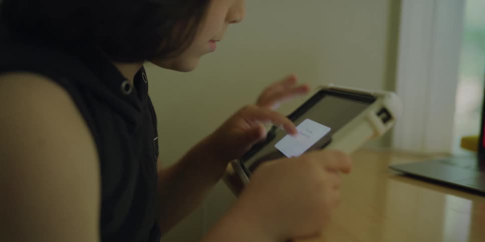 Little girl with dark hair and a sleeveless shirt sits at table and uses a tablet in a room