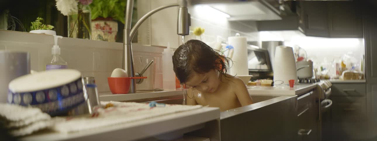 A small child sits in a kitchen sink with the faucet running water over their head