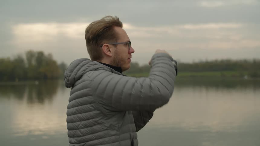 A young man at a lake turns and takes a picture with his camera