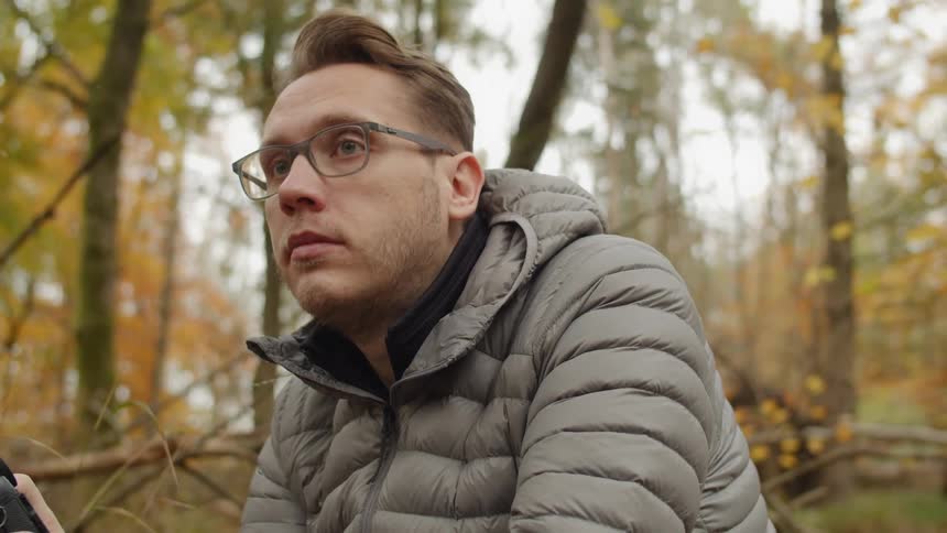 A man wearing glasses and a gray coat sits in the forest taking photographs with a camera