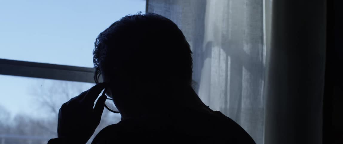 A man stands in a room looking out of a window while he puts on a pair of glasses
