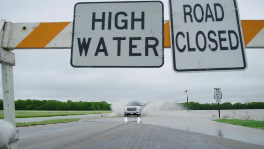 A vehicle driving through deep water on a road stops at a road closed sign spraying flooded water to both sides of the vehicle