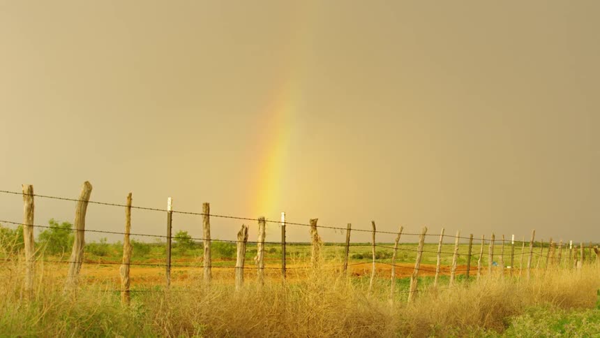 A rainbow in the sky on a day with overcast in the sky behind a fence make of barbed wire and sticks