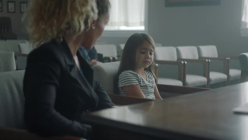 A woman speaks to a little girl while sitting in chairs behind a table with empty row of chairs in the same room