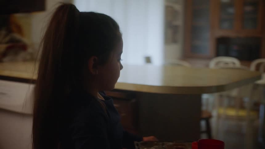A young girl retrieves a tray of food from a kitchen counter and carries it through the kitchen and dining room towards a corridor