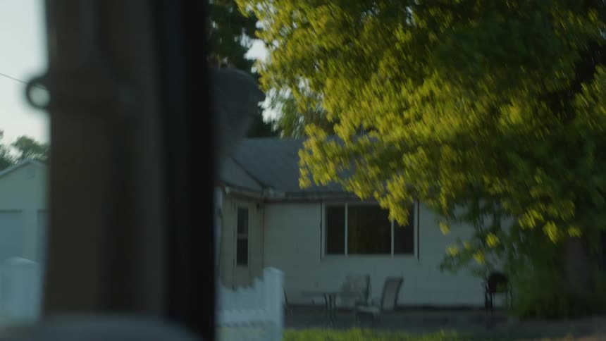 A vehicle drives with its back window down past a small white house in a neighborhood