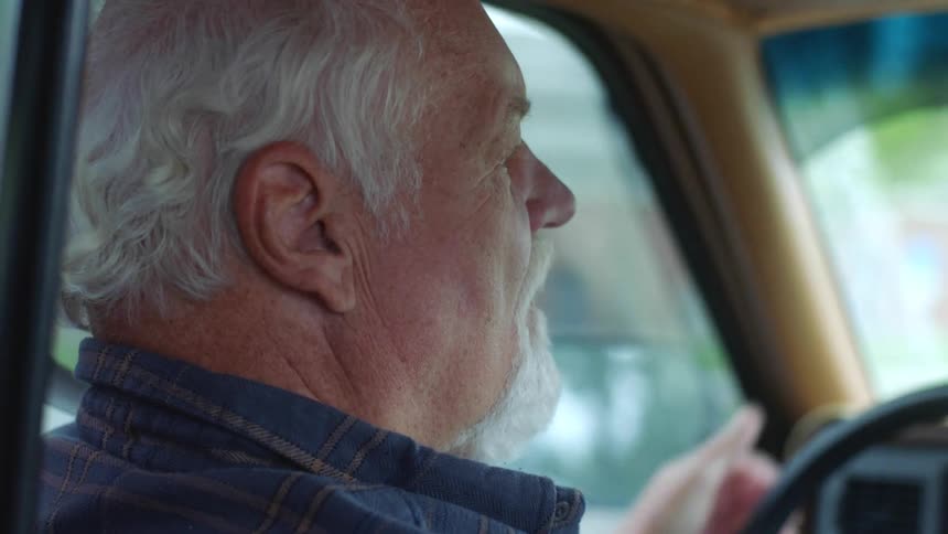 An older man with white and a beard carefully turns the steering wheel in his car