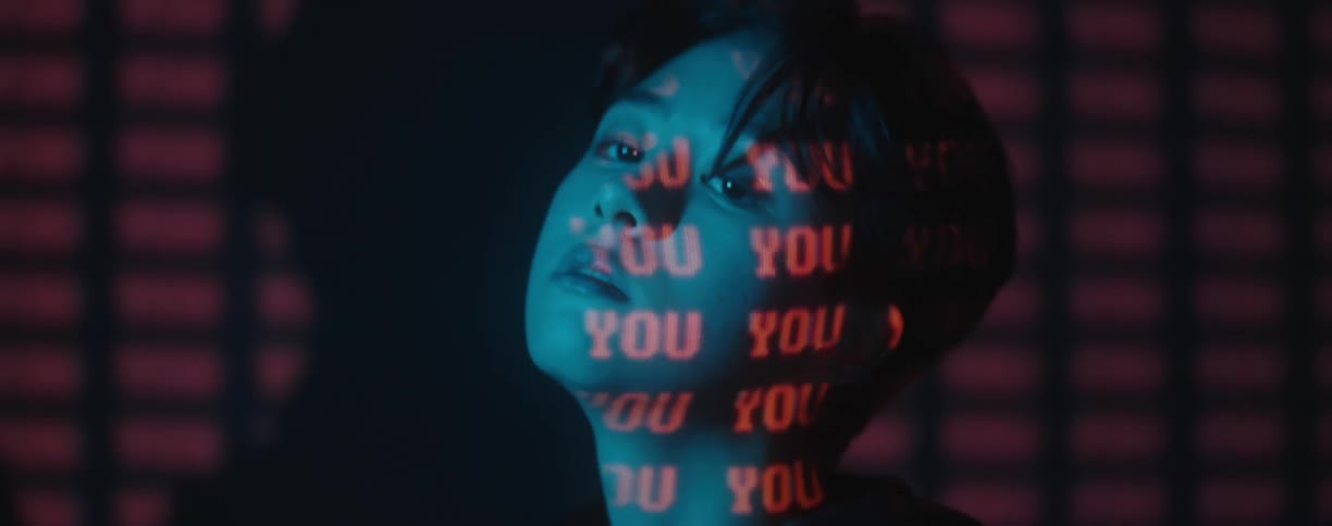 Woman with head laying on her arm raises her head with the word you projected in red on her face neck and the wall behind her