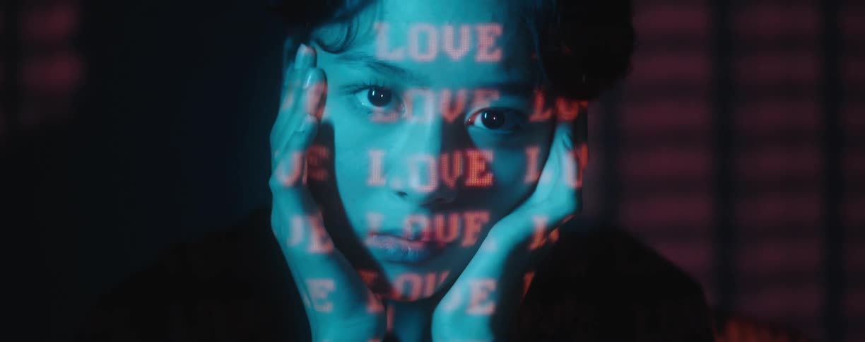 Woman sits rubbing face and neck with hands while the word love is projected in red on her