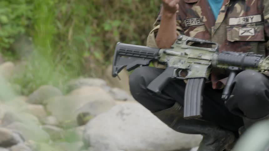 A man with an assault rifle squats near a stream