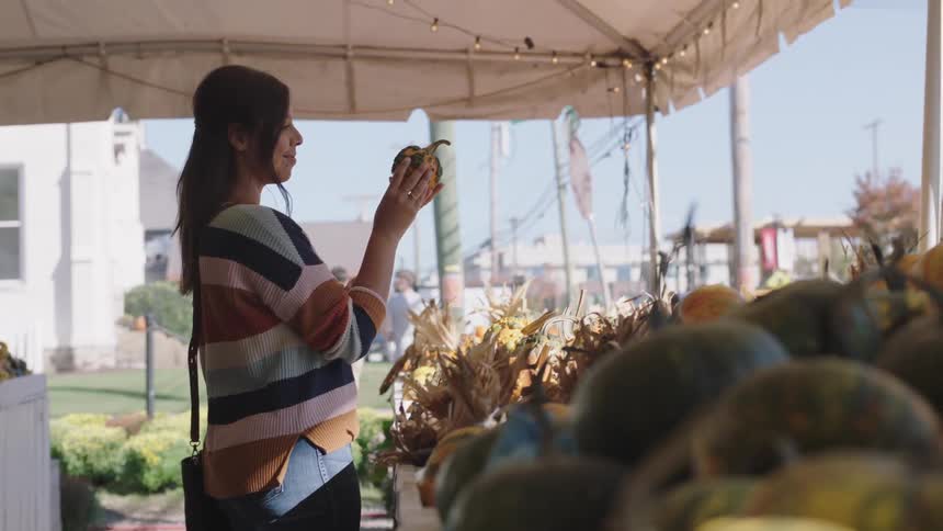 A woman in a striped sweater looking at a small pumpkin at a produce stand