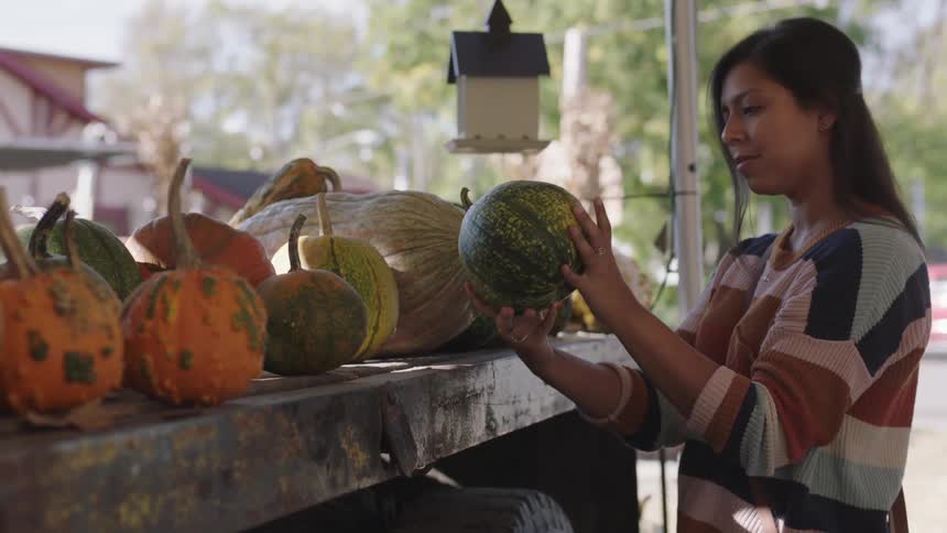 A woman in a sweater picks up a large squash and inspects it