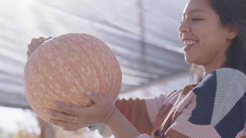 A woman smiling while holding a pumpkin outside on a sunny autumn day