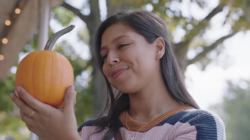 A woman standing outside of a building examines a small pumpkin before turning to smile into the camera