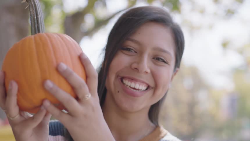 Brunette woman holds a small pumpkin and smiles at the camera