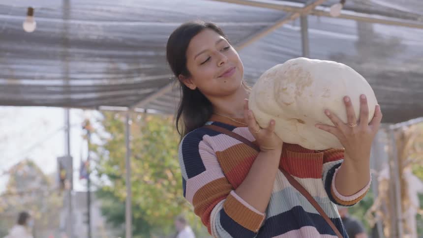 A woman examines a squash at an outdoor market on a busy street corner and places it in a wagon
