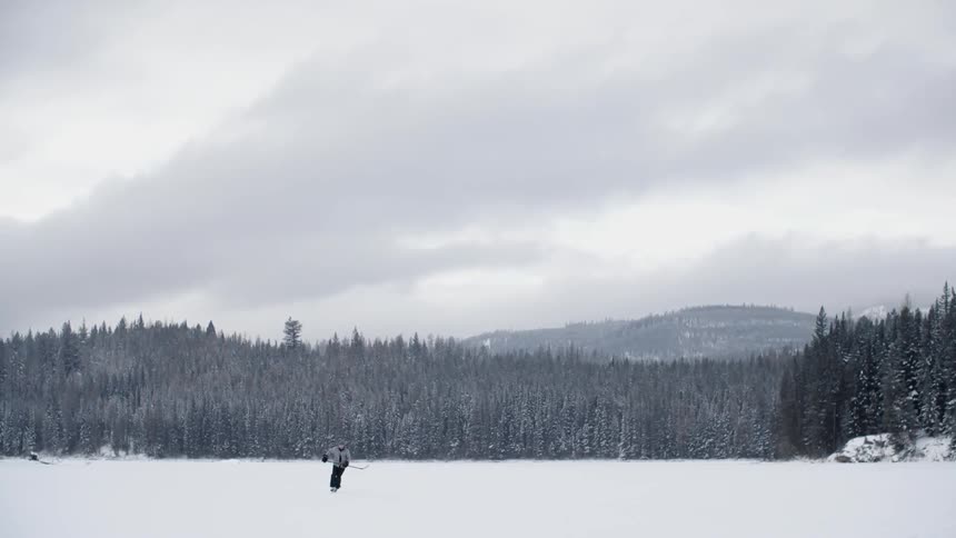 A person skates across a snowy ice rink holding a hockey stick