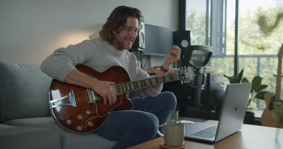 Man sits on couch holding guitar talking and laughing to someone on video call via laptop