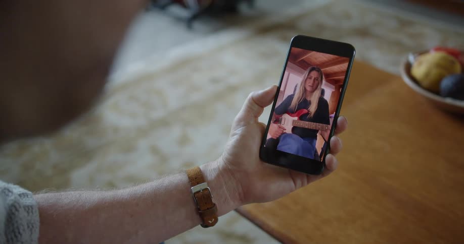 A man sitting in a living room uses a smartphone to video chat with a woman playing the guitar