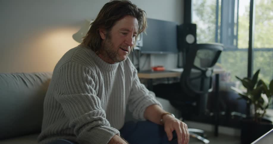 Man with long hair and a grey pullover opens the laptop to make a video call in the living room at home