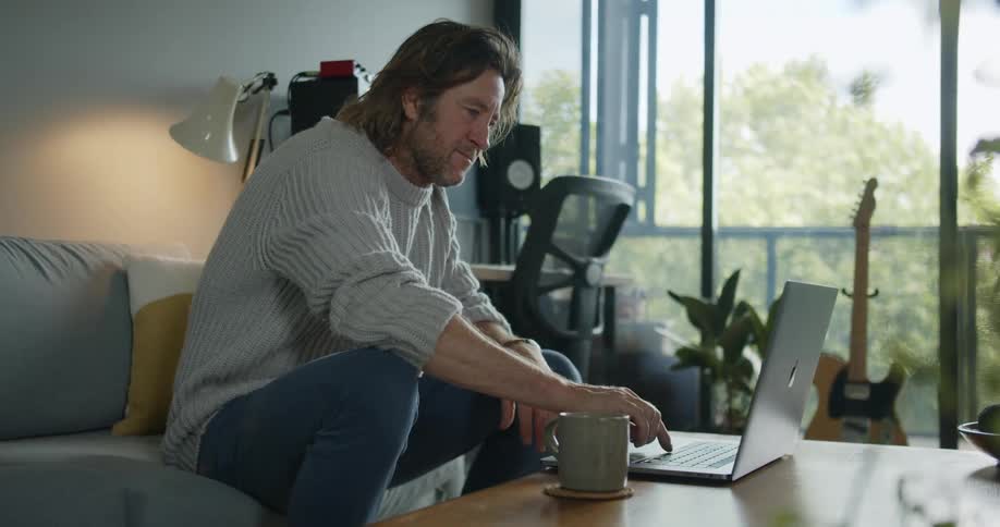 A middle aged man sitting on a sofa in a home living room opens a laptop and smiles at the screen