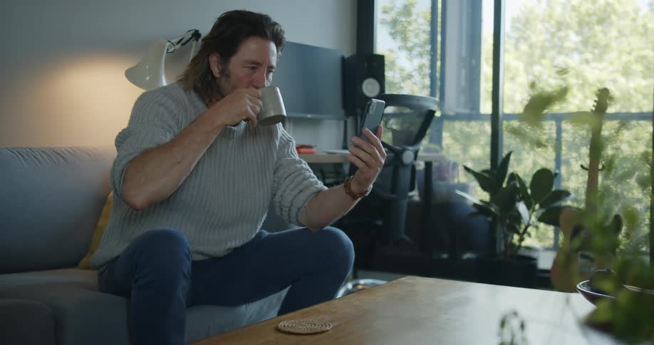 Man sitting in a couch at home talks on the phone while drinking a hot beverage