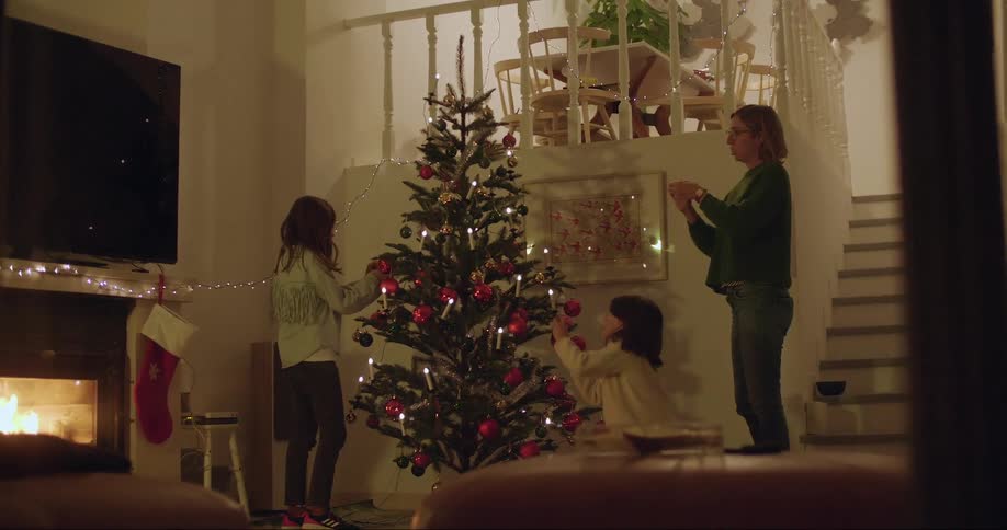 Mom and two daughters decorate a christmas tree in the living room with a fire in the fireplace at night