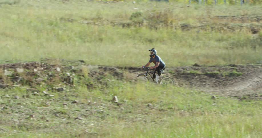 A man rides a bike through a forest exiting on a ramp and moving over a dirt path with a hill during the day