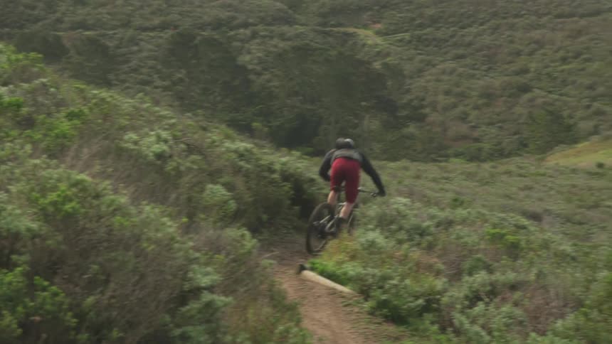 A cyclist rides a bicycle at high speed along a narrow dirt path through a hilly countryside during the day