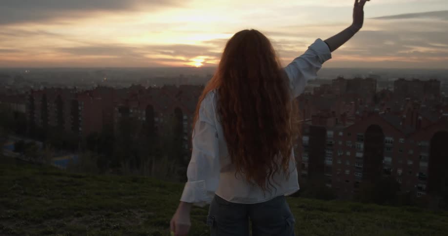 A woman stands on a hill above a neighborhood and stretches her arms in the air