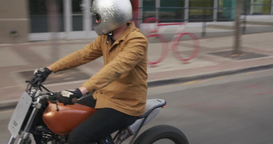 A man in a tan jacket rides a small motorbike through a city