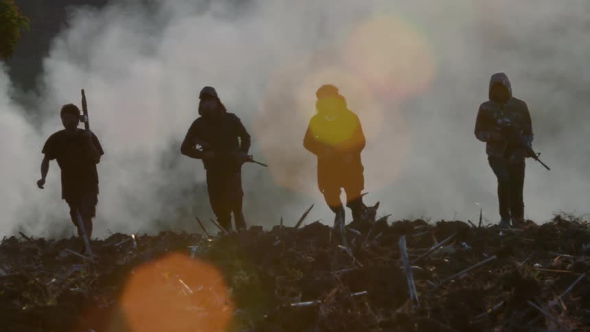 Armed men walking across a field with smoke behind them and their dog following